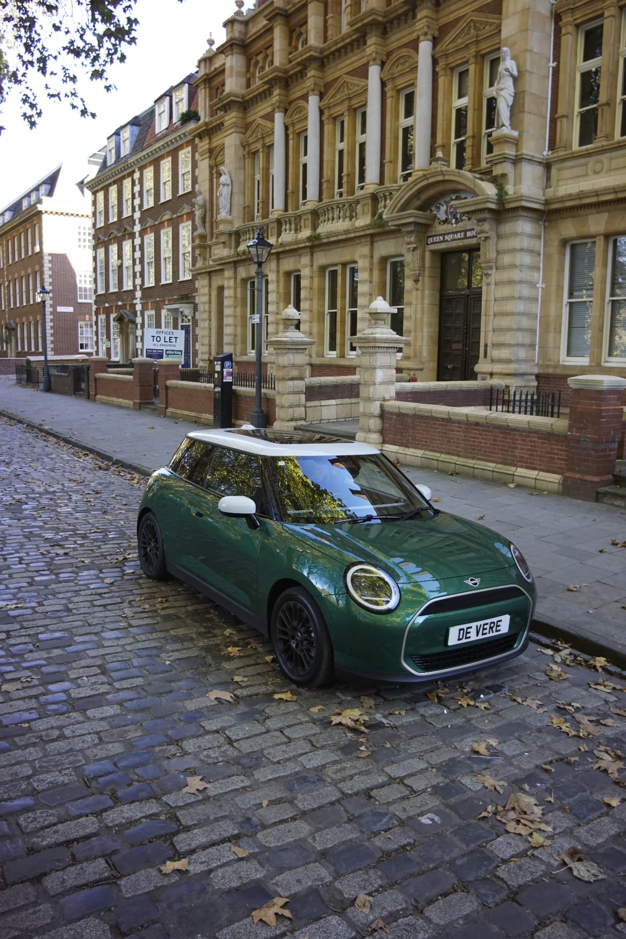 Green MINI Cooper E with white roof and circular LED headlights on a cobbled street